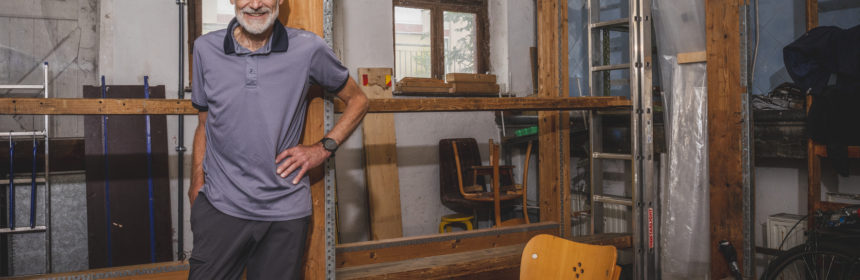 Wolrad Specht trägt ein graues Polohemd und eine dunkelgraue Hose. Er steht in der vorderen linken Hälfte des Bildes. Im Hintergrund ist der Rahmen zu sehen, auf dem er die Teppiche aufspatnnte.