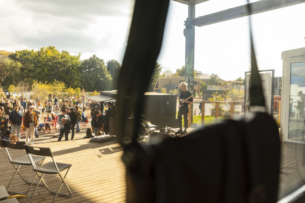 SCHRAMM, ein Wuppertal Musiker steht in der rechten Bildhälfte mit seiner Gitarre. In der Linken Bildhälfte ist das Publikum zu sehen. Das Bild ist durch einen Gegenstand im Vordergrund geteilt